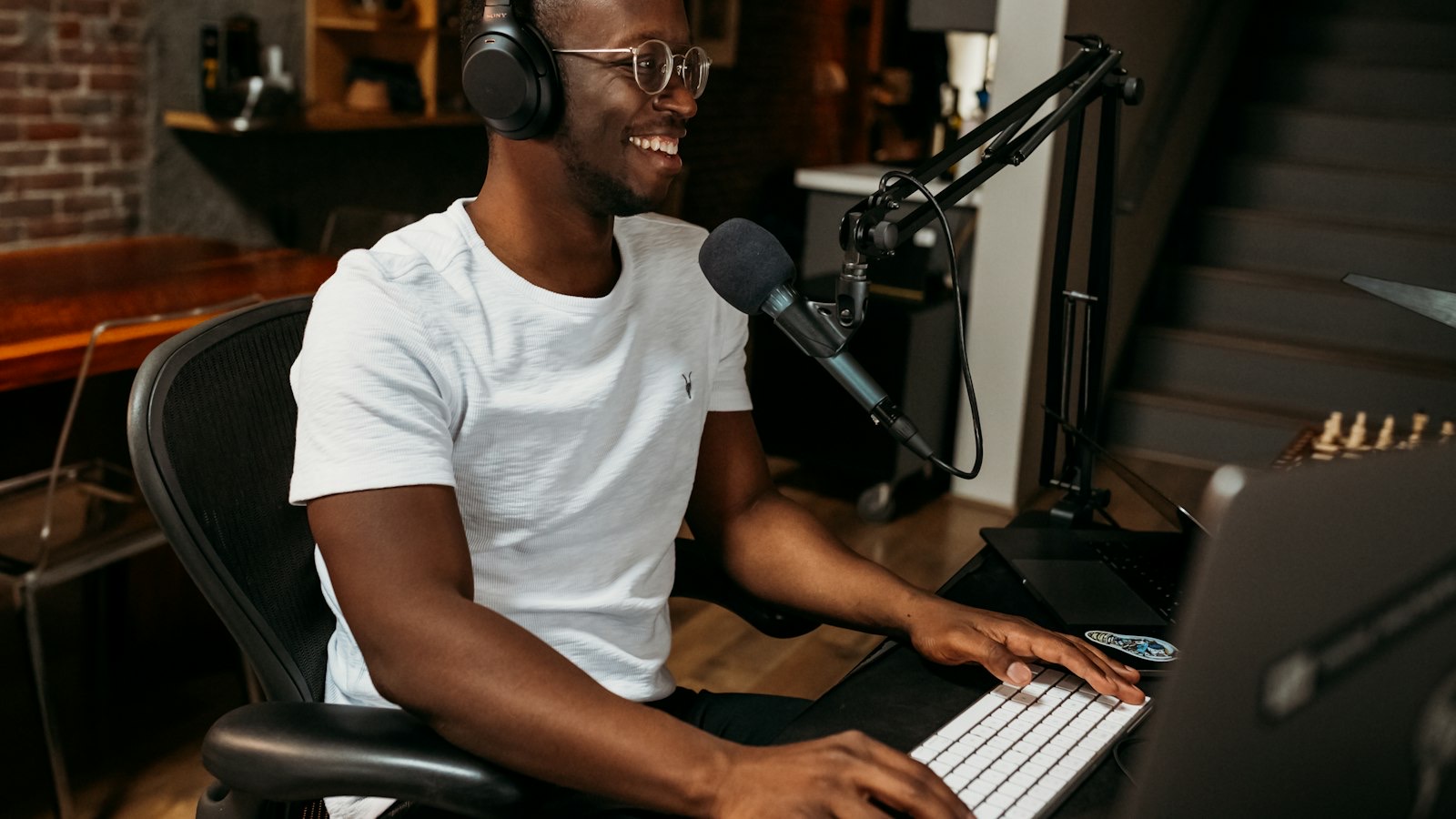 A presenter in headphones smiling into a microphone at a desk with a laptop