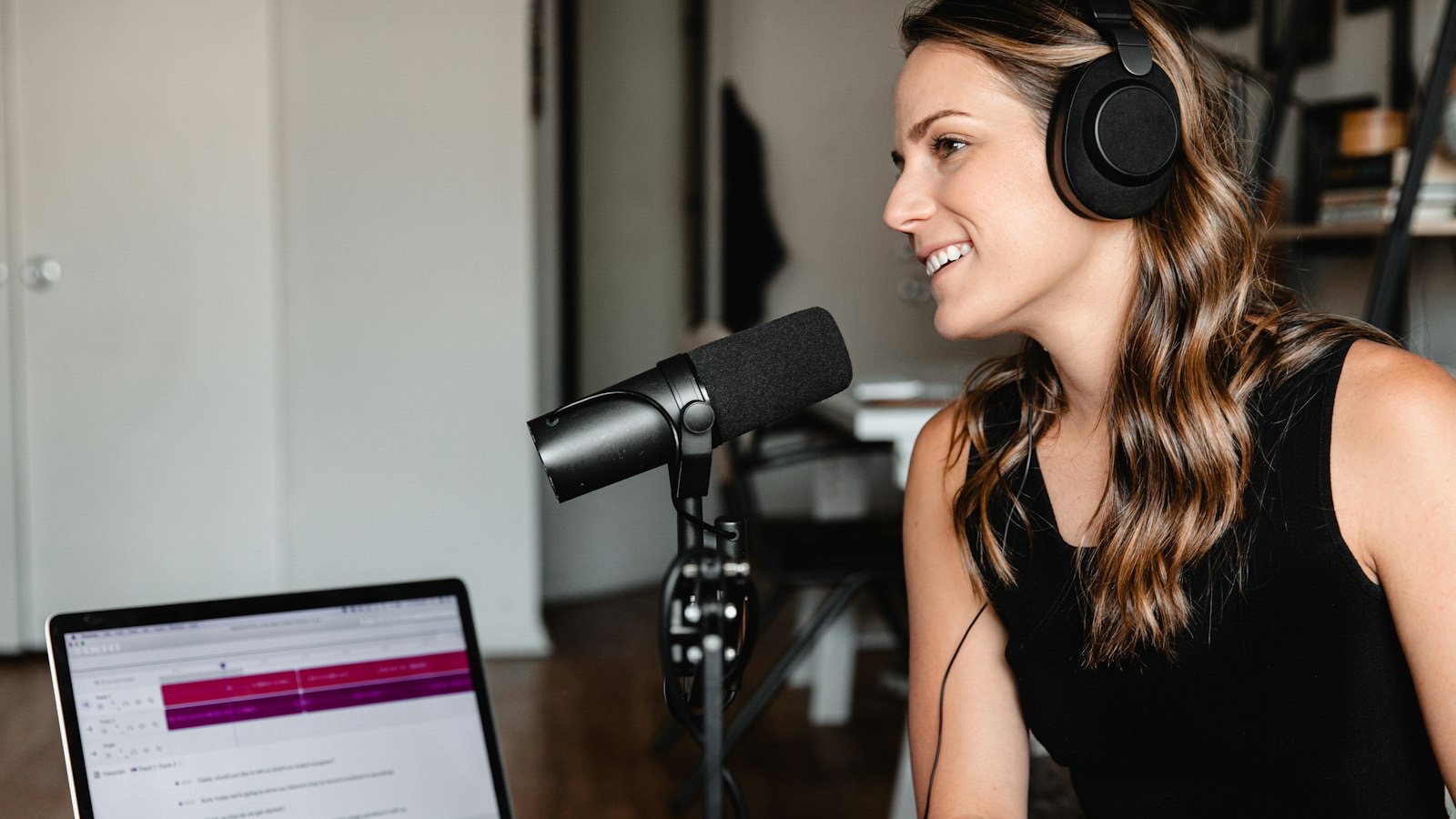 A podcaster in headphones talking into a microphone beside a laptop showing a recording