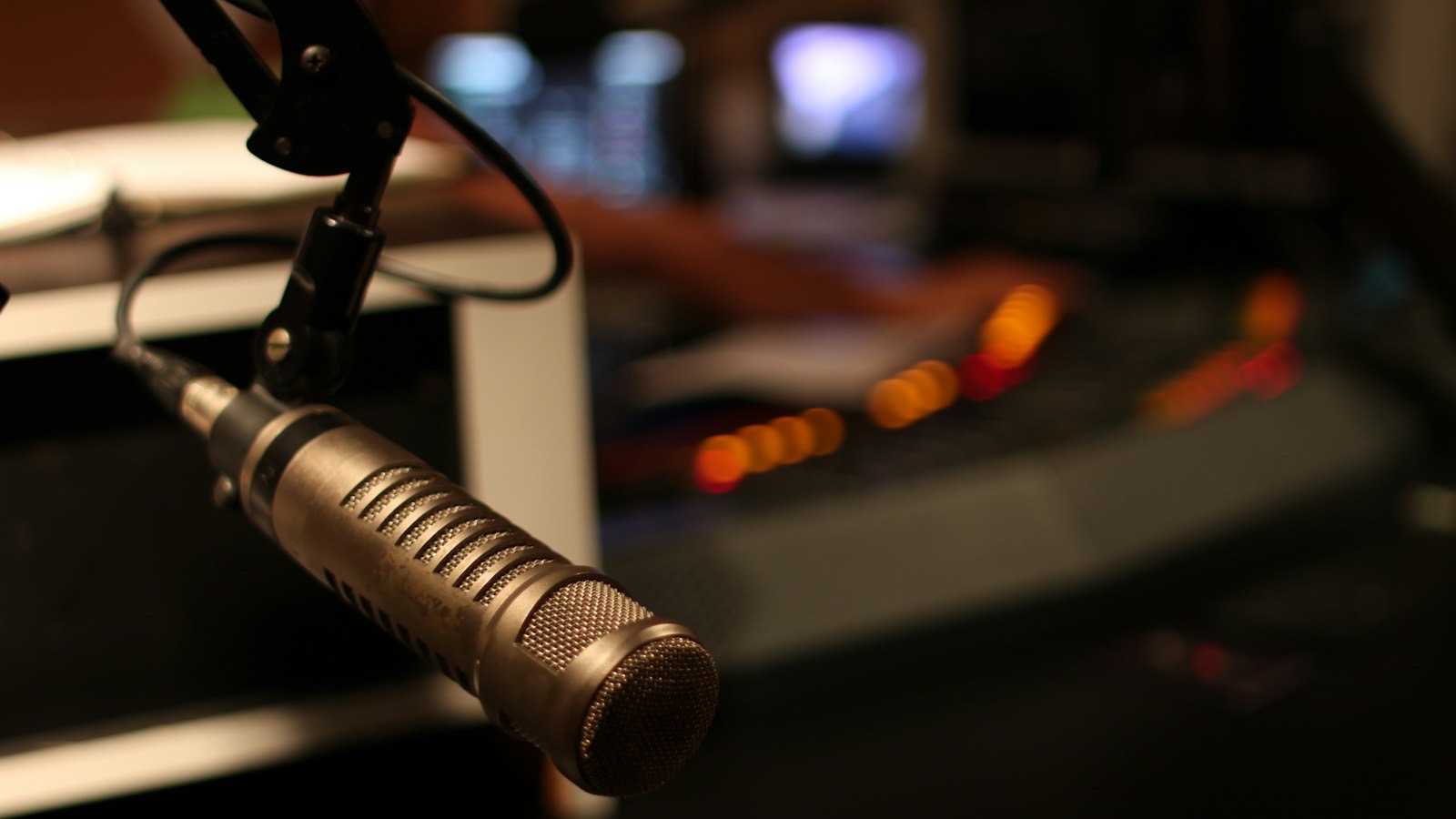 A broadcast microphone on a boom arm above a lit studio mixing desk